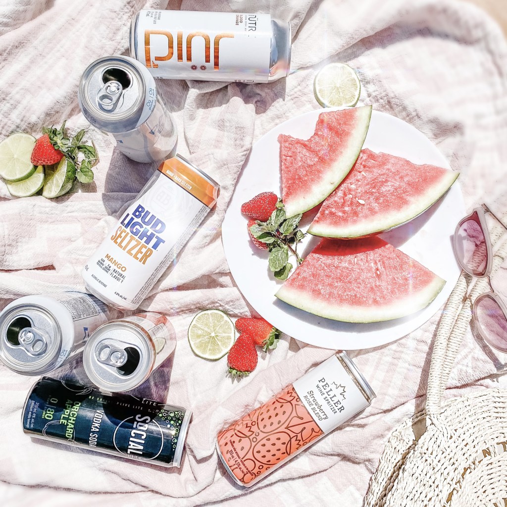 Low-Calories Summer Alcoholic Coolers laid on a picnic blanket with fresh fruit. Low-Calorie 2021 Summer Alcoholic Drink Review: The best low-sugar alcoholic coolers available in stores now at the LCBO.